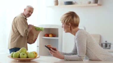 Bir kadın mutfak adasında otururken akıllı telefonuna odaklanır. Ortağı, arkasındaki kese kağıdından poşetleri açıyor. Taze meyveler yakınlarda sergilenir..