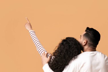 Rear view of young arab couple embracing and pointing up while sitting on floor over yellow studio background together, millennial brunette woman demonstrating copy space for advertisement, panorama clipart