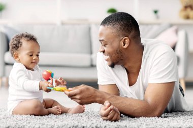 Bebeklerle Gelişim Faaliyetleri. Genç siyahi babanın evde bebekle oynadığı portresi, mutlu Afrikalı Amerikalı adam sevimli bebeğine oyuncak veriyor ve gülümsüyor.