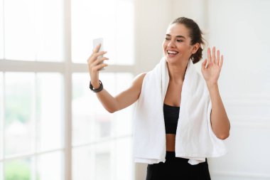 Havlusu olan atletik genç bir kadın spor eğitmeniyle online konferans yapıyor ya da video blogu kaydediyor. Sportif güzel bayan akıllı telefon ekranına el sallıyor, eğitmenini veya takipçilerini selamlıyor, boş alan.