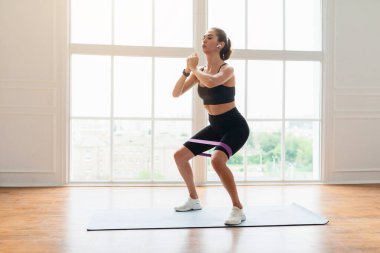 Eğitim Konsepti. Kendinden emin sporcu kadın fitness stüdyosunda elastik direnç bandıyla ya da oturma odasında bacak ve kalça egzersizi yaparak kulaklıkla müzik dinleyerek çömeliyor.
