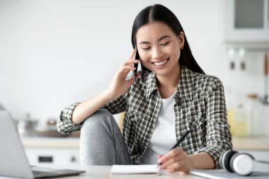 Gülümseyen Asyalı serbest çalışan genç bir kadın cep telefonuyla konuşuyor ve not alıyor, izolasyon sırasında evden çalışıyor, kopyalama alanında çalışıyor. Mutlu bayan telefonda konuşuyor, mutfakta dizüstü bilgisayar kullanıyor.