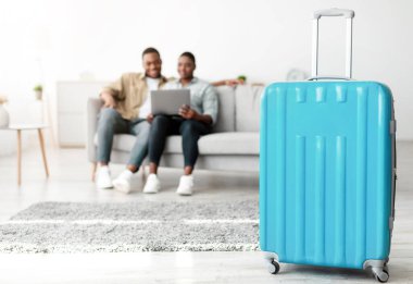 Aile Tatili Konsepti. Mavi bavul, tanınmayan Afro-Amerikan çift evdeki koltukta dizüstü bilgisayar kullanırken yerde duruyor. Bavula Seçici Odaklanma