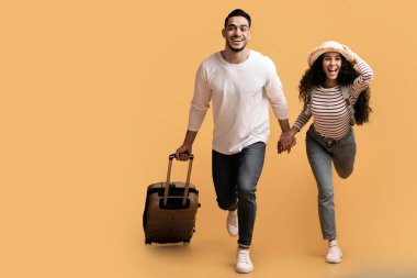 Tatile hazırım. Sarı arka planda koşan neşeli Arap çift, Orta Doğu 'lu mutlu eşlerin portresi el ele tutuşuyor ve valiz taşıyor, tam uzunluk, kopyalama alanı.