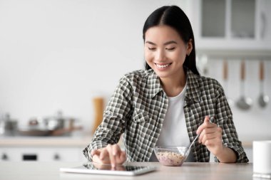 Gülümseyen Asyalı kadın kahvaltı yapıyor ve sosyal medyada sörf yapıyor, dijital tablet, mutfak içi, fotokopi alanı kullanıyor. Yulaf ezmesi yiyip blog ya da kitap okuyan mutlu genç bayan.