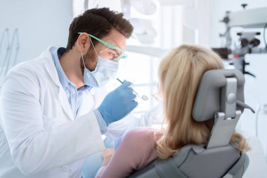 Yüzü maskeli ve gözlüklü bir erkek hasta sarışın kadın için tedavi görüyor, diş aletleri tutuyor, lastik eldiven takıyor. Stomatoloji, dişçilik, modern diş sağlığı kliniği konsepti