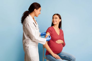 Covid-19 Aşılama. Coronavirüs 'e karşı aşı olan hamile kadın hasta, Aşılanmış Koluna Mavi Stüdyo Arkaplanı Üzerine Alçı Bandajı uygulayan Tıp işçisi