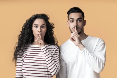 Young arab man and woman showing shh gesture at camera, mysterious middle eastern couple keeping forefingers on lips, making silence sign while standing together over yellow background, copy space clipart