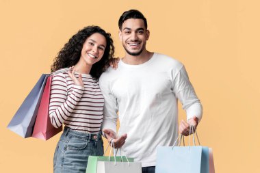 Sarı arka planda alışveriş torbaları olan neşeli, genç Orta Doğulu çift pozları, Alış-veriş yapan mutlu bir Arap ve kadın, sezonluk satışların tadını çıkarıyor, kopyalama alanı.