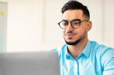 Uzaktan çalışma konsepti. Laptop kullanan yakışıklı bir Arap modern ofisteki internette bilgi arıyor. Odaklanmış Doğulu girişimci internet üzerinden iş görüşmesi yapıyor, çalışanlarla konferans yapıyor.