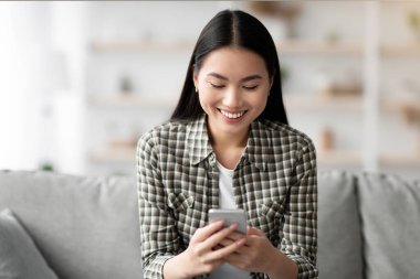 Akıllı telefonuyla koltukta oturan mutlu Asyalı kadın, sohbet ediyor ya da e-postaları kontrol ediyor, ev içi, fotokopi odası. Gıcır gıcır cep telefonu kullanan, sosyal medyayı kontrol eden ve gülümseyen genç bir bayan.