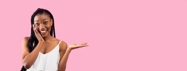 Yüzüne dokunan şık elbiseli heyecanlı Afro kadın, pembe stüdyo arka planında boş bir alanda bir şeyler gösteriyor, afiş. Çekici siyahi bayan ürününüzün veya hizmetinizin reklamını yapıyor.