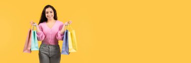 Mevsimlik satış konsepti. Bir sürü renkli alışveriş çantası taşıyan, sarı stüdyo arka planındaki kameraya gülümseyen, günlük kıyafetli, neşeli genç bir kadın.