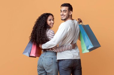 Aile alışverişi. Mutlu Arap eşler renkli alışveriş çantalarını kucaklıyor ve tutuyor, sarı stüdyoda yürüyen gülümseyen orta doğulu çift ve kameraya doğru dönüyorlar, tam uzunlukta, kopyalama alanı.