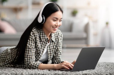 Evde tek başına dinlenirken kablosuz kulaklık ve laptop kullanan neşeli Asyalı kadın oturma odasında halıya uzanmış video oyunları oynuyor ya da internetten film izliyor. Ev eğlencesi