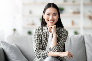 Evdeki kanepede oturan, kameraya gülümseyen, uzun saçlı Asyalı mutlu bir kadın. Ev dekorasyonu, milenyum yaşam tarzı, rahatlama ve rahatlama kavramı.