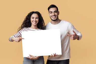 Reklam için yer. Ellerinde boş beyaz plaketi tutan ve gülümseyen Orta Doğulu genç erkek ve kadın reklam için kopya alanı gösteren mutlu bir Arap çifti.
