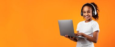 Çocuklar ve Teknoloji Konsepti. Gülümseyen siyah kızın portresi dizüstü bilgisayar kullanıyor, kablosuz kulaklık takıyor, çalışıyor, çevrimiçi kurslar izliyor, sarı stüdyo duvarından izole edilmiş.