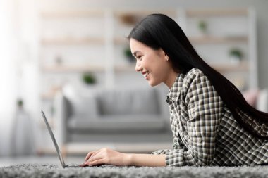 Evde dizüstü bilgisayar kullanan Asyalı genç bir kadının yan görüntüsü, oturma odasında halıya uzanması, bilgisayar klavyesine yazı yazması, hafta sonları dinlenirken sevgilisiyle veya arkadaşlarıyla sohbet etmesi, uzay kopyası.