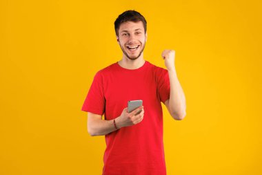 Büyük Galibiyet İnternette. Gülümseyen Gündelik Genç Adam 'ın Portresi Cep Telefonu Kullanarak Başarı Kutlaması. Mutlu Adam Kazanan Jest Yapıyor, Gadget 'ı tutuyor ve sıkılmış yumruğunu sallıyor, Mavi Stüdyo' da poz veriyor.