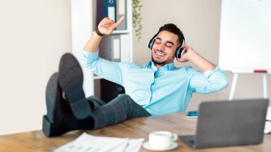 Ofiste mola veriyorum. Neşeli bir Arap kablosuz kulaklıkla müzik dinliyor, iş yerinde dinleniyor. Pozitif genç adam dizüstü bilgisayarla masada oturuyor ve favori şarkının tadını çıkarıyor.