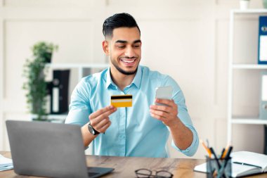 İnternetten alışveriş konsepti. Kredi kartı ve akıllı telefon tutan mutlu Arap işadamı e-ödeme yapıyor, internetten mal sipariş ediyor, ofisinden bir şeyler satın alıyor.