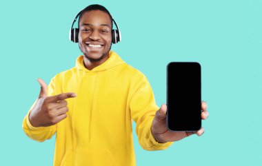 Müzik Uygulaması. Neşeli Siyah Adam, Mavi Stüdyo Arkaplanı Üzerine Gülümseyen, Kablosuz kulaklık takan, Boş Telefon Ekranı Gösteriyor. Uygulama Reklam Modeli. Seçici Odaklanma