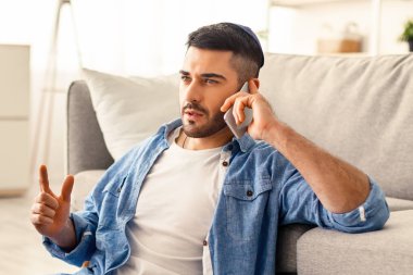 Kurumsal İş İletişim Konsepti. Odaklanmış genç Yahudi bir adamın portresi telefon görüşmesi yapıyor, bilgisayarda çalışıyor, yerde oturuyor ve oturma odasında kanepeye yaslanıyor, proje planını tartışıyor.