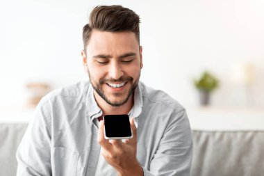 Mutlu yakışıklı adam akıllı telefondan sesli mesaj kaydediyor ya da evdeki kanepede dinlenirken sanal asistan kullanıyor. Gülümseyen adam iletişim ve serbest alan için akıllı teknolojilerden zevk alıyor.