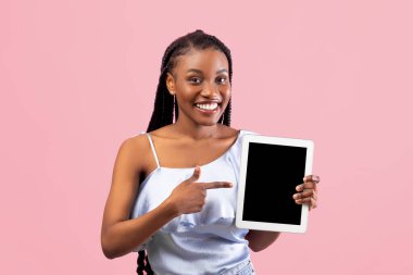 Pozitif genç Afro kadın pembe stüdyo arka planında boş ekranı olan tablet bilgisayarını işaret ediyor, web sitesi ya da uygulama için model alıyor. Havalı siyahi bayan reklam için boşluk sunuyor.