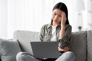 Dizüstü bilgisayar kullanırken başı ağrıyan yorgun Koreli kadın, kanepede oturuyor ve evde çalışıyor, kafasına dokunuyor, kopyalama alanı yaratıyor. Genç Asyalı kadının bilgisayarla ilgili sorunları var.