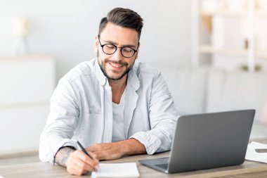 Mutlu milenyum adamı online iş arıyor, masada oturuyor, dizüstü bilgisayar kullanıyor ve notlar alıyor, içerisi oturma odası, boş yer. Evden çalışan heyecanlı bir adam, uzaktan kumandalı.