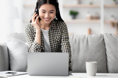 Genç Asyalı olumlu bir kadın evinden bilgisayar ve kulaklık kullanarak çevrimiçi konferans veriyor bilgisayar ekranına bakıyor ve gülümsüyor. Webinar 'a katılan neşeli esmer bayan