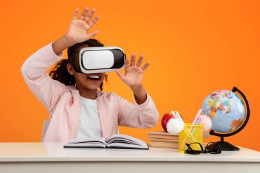 Sanal Gerçeklik Konsepti. VR kulaklıklı heyecanlı Afrikalı Amerikalı kızın portresi sarı stüdyoda, fotokopi alanında havaya dokunuyor. Masada oturmuş yapay bilgisayar dünyasını keşfeden pozitif bir genç.