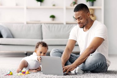 Bebekle Serbest Çalışmak. Genç Afrikalı bir adam dizüstü bilgisayarda çalışıyor ve bebeğine evde bakıyor, Kara Baba bilgisayarını online iş için kullanıyor ve bebeğe bakıcılık yapıyor.