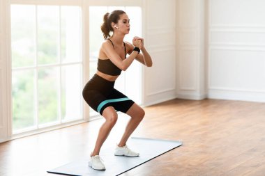 Body Shaping And Glutes Workout. Determined Fit Young Woman In Black Sportswear Exercising With Resistance Loop Band Doing Squats Standing On Yoga Mat, Training Butt And Hamstring, Wearing Earbuds
