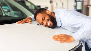 Mutlu Afrikalı Amerikalı Adam, yeni arabasının kapüşonlusunu kucaklıyor, neşeli gülümseyen milenyum siyahi adam, araba satın almayı hayal ediyor, lüks otomobilin yüzeyine yaslanıyor, yakın plan,