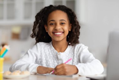 Smiling black girl studying online, using laptop at home, taking notes or drawing, looking ar camera and smiling. Cute african american kid having online lesson, kitchen interior. Home schooling clipart