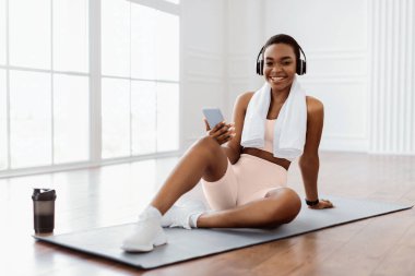 Kablosuz kulaklıklı, akıllı telefonlu, spor kulaklıklı, spor ayakkabılı, spor salonunda çalışan, su içen, kameraya bakan ve gülümseyen, bedava fotokopi alanı olan formda bir bayan.
