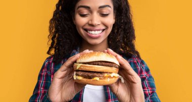 Abur cubur. Biftek ve peynirli hamburger tutan komik, Afro-Amerikan kadının yakın plan portresi. Kıvırcık saçlı, lezzetli sandviçi yemeye hazır, turuncu arka planda izole edilmiş siyah kadın, afiş.