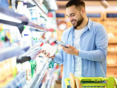 Gülümseyen genç adam marketteki raflardan süt ürünleri alıyor, elinde şişe ve akıllı telefon, müşteriler barkodu tarıyor cep telefonuyla, el arabası ile yürüyor.