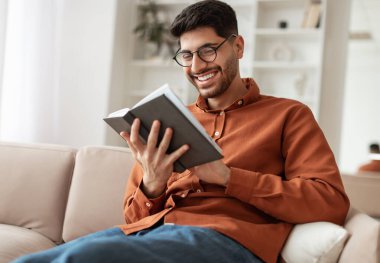 Dinlenme ve gevşeme kavramı. Gözlük takan neşeli Orta Doğulu genç bir adamın portresi. Elinde kağıt kitap, evinde kanepede oturuyor, edebiyat okuyor, hikayeden zevk alıyor.