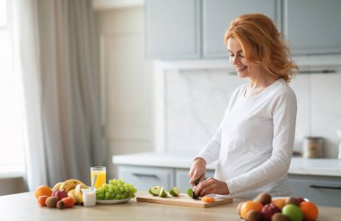Hamile bir kadın renkli meyvelerle dolu parlak bir mutfakta elma ve üzüm gibi taze meyve dilimliyor. Sağlıklı bir sabah atıştırması hazırlarken gülümsüyor. Yanında bir bardak meyve suyu ile, boşluğu kopyalıyor.