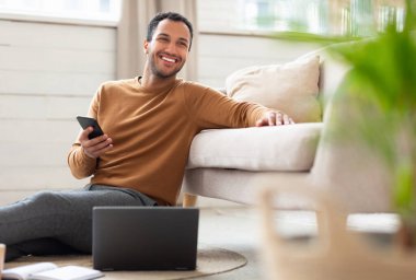 Serbest meslek hayatı. İç İşleri 'nde çalışan heyecanlı Orta Doğulu erkek cep telefonunun portresi, bilgisayar kullanan, oturma odasında kanepeye yaslanan, başka yere bakan mutlu adam.