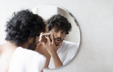 Problem skin. Concerned indian man popping pimple on cheek while standing near mirror in bathroom. Unhappy guy inspecting face, suffering acne, focus on reflection