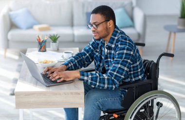 Engelliler için internet işi. Tekerlekli sandalyedeki özürlü adam evdeki uzaktan kumanda için dizüstü bilgisayar kullanıyor. Milenyum belden aşağısı felçli bir adam internette iş görüşmesi yapıyor, video görüşmesi yapıyor.