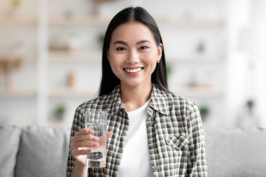 Oturma odasında kanepede otururken elinde tatlı suyla gülümseyen Asyalı kadın, pozitif genç kadın günlük su içmeyi öneriyor, sağlıklı bir yaşam tarzı var, yer kopyası.