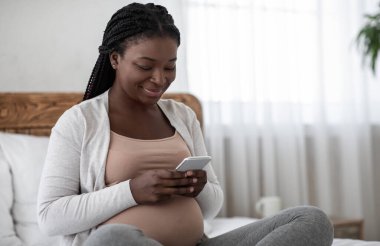 Hamilelik Takip Uygulaması. Mutlu siyahi hamile kadın evde akıllı telefon kullanıyor, gülümseyen anne bebek gelişimi hakkında bilgi okuyor, cep telefonuyla yatakta oturuyor, yatak odasında dinleniyor.