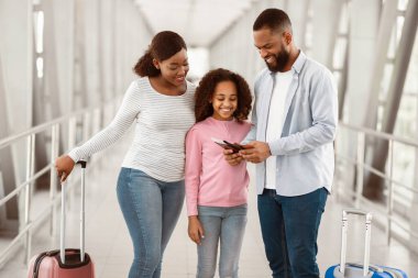 Modern havaalanı terminalinde bavullarıyla bekleyen üç kişinin neşeli siyah aile portresi, baba elinde pasaport, anne ve kız kucaklaşması,