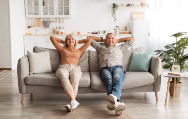Happy Mature Couple Sitting On Couch Relaxing And Resting At Home, Holding Hands Behind Head, Enjoying Comfort Of Their House. Relaxed Senior Spouses Having Lazy Weekend Indoors. Full Length